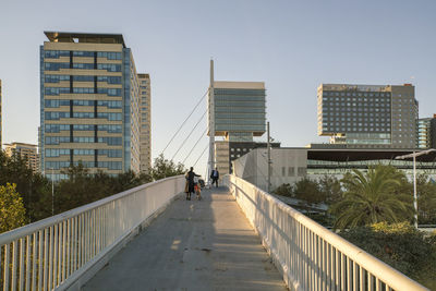 People on footbridge amidst buildings in city against clear sky