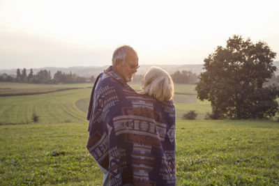 Happy senior couple standing on a meadow at sunset sharing blanket