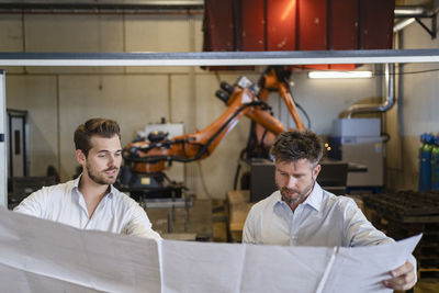 Businessmen holding blueprint paper while standing against automated machine at factory