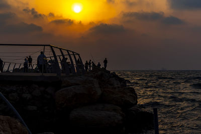 People on rocks by sea against sky during sunset