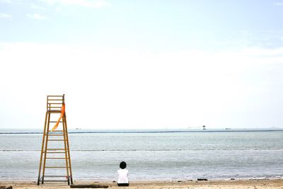 Rear view of woman sitting at beach against sky