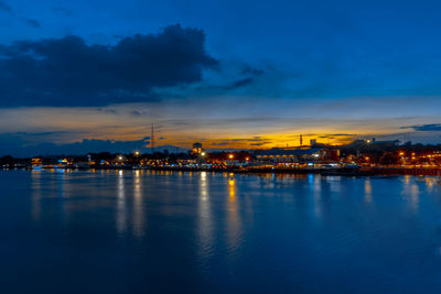 Illuminated city by sea against sky at sunset