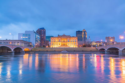 Illuminated bridge over river by buildings against sky at dusk