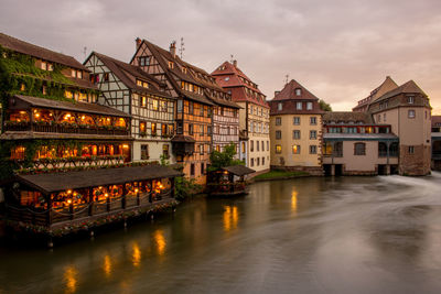 Illuminated buildings by river against sky in city at dusk