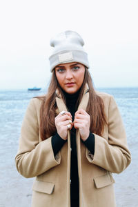 Portrait of beautiful woman in warm clothing standing against sea