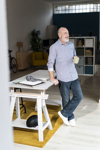 Smiling senior freelancer holding coffee mug standing by desk at home