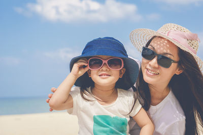 Happy mother and daughter wearing sunglasses enjoying at beach against sky