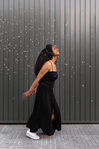 Joyful african american female with curly hair scattering white confetti in city while having fun and looking up