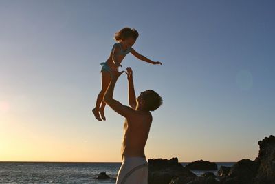 Happy father throwing daughter in air at beach against clear sky during sunset