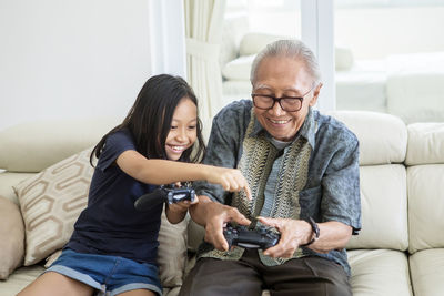 Happy senior man with granddaughter playing video game while sitting on sofa at home