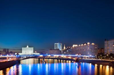 Illuminated bridge over river by buildings against blue sky at night