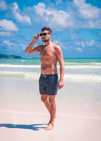 Full length of shirtless man in sunglasses walking at beach against sky