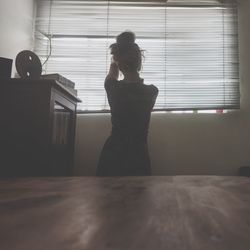 Rear view of woman standing against window at home