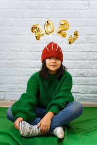 Funny asian girl in green sweater, sits on floor with golden balloons with number 2023 in a red hat