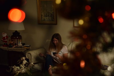 Smiling woman using mobile while sitting on sofa at home during christmas
