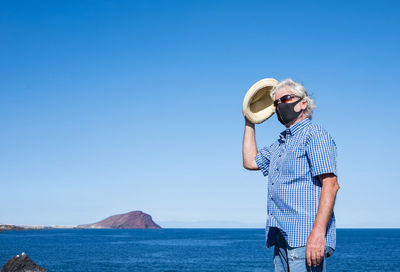 Man with face mask standing by sea against clear blue sky