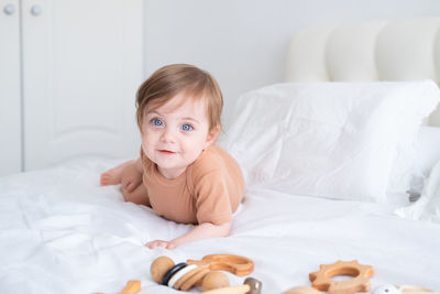 Baby girl with blue eyes in brown bodysuit playing with wooden toys on white bedding on bed