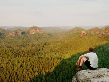 Moment of loneliness. tourist in shirt sit on the peak of rock and watching into forest valley.