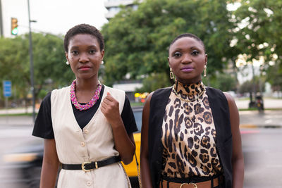 Feminine adult black women with short dark hair in fashionable outfits and accessories standing together on city street and looking at camera