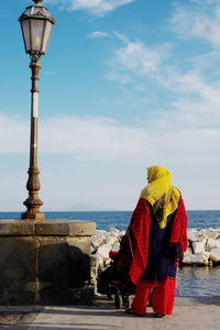 Rear view of mid adult woman in traditional clothing standing by sea on footpath against sky