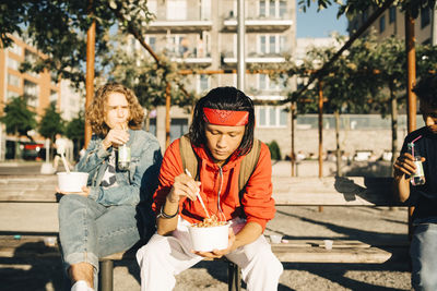 Friends eating meal from containers while sitting on bench in city during sunny day