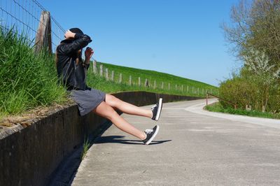 Side view of woman sitting on retaining wall by road against sky during sunny day
