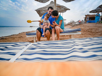 Family sitting on lounge chair at beach against sky