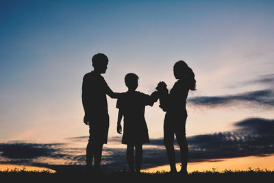 Low angle view of silhouette friends with puppy standing against sky during sunset