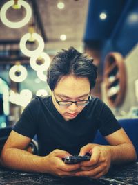 Young asian man in eyeglasses using smartphone at table in cafe against ceiling led lights