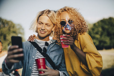Smiling man taking selfie with friend on smart phone while enjoying drinks in concert