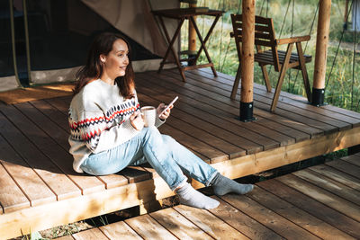 Brunette woman in nordic sweater using mobile phone and relaxing in glamping in nature