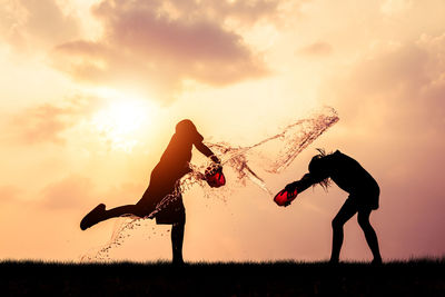 Full length silhouette friends splashing water on each other while standing against sky during sunset