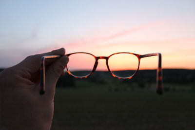 Close-up of human hand holding eyeglasses against sky during sunset