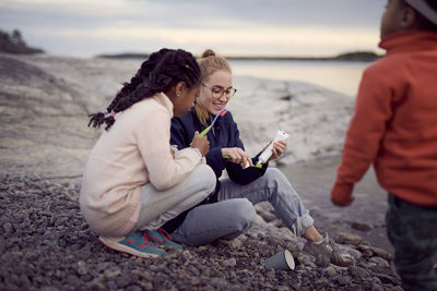 Smiling mother removing toothpaste on toothbrush while sitting by daughter at beach