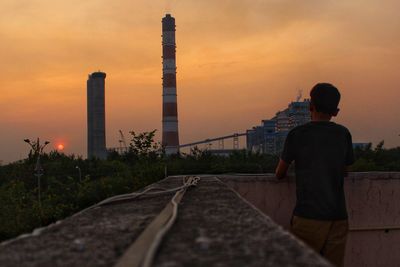 Rear view of man standing on in front of industy against sky during sunset