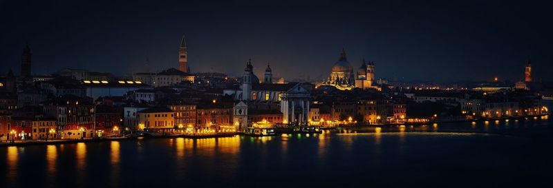 Illuminated buildings in front of river against sky at night