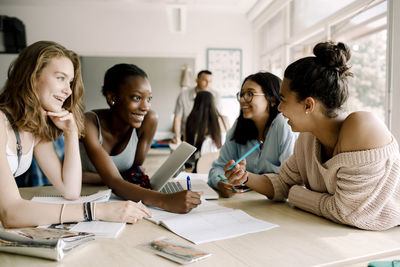 Smiling female teenagers discussing while sitting by table in classroom