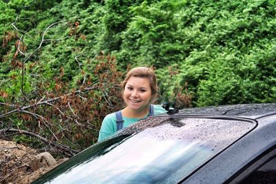 Portrait of smiling young woman by car against trees