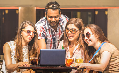 Cheerful friends using laptop on table at outdoor cafe during sunny day