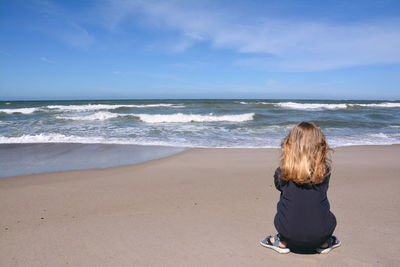 Rear view of girl crouching on shore at beach against sky