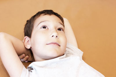 Close-up of thoughtful boy with hands behind head against brown wall