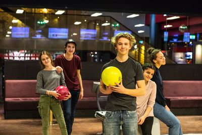 Portrait of smiling teenage boy holding ball while standing against friends at bowling alley