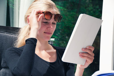 Smiling beautiful young woman holding sunglasses while using digital tablet outside house