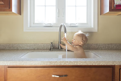 Shirtless baby boy playing with water while sitting in kitchen sink