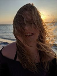 Close-up of woman with tousled hair at beach against sky during sunset