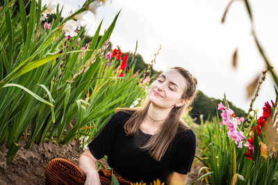 Smiling young woman with eyes closed sitting by flowering plants on land