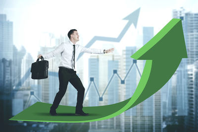 Digital composite image of smiling businessman with bag standing on green arrow symbol in city