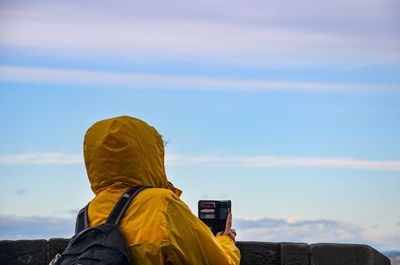 Rear view of person in yellow raincoat photographing against sky