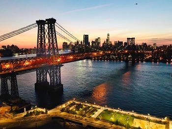 Illuminated bridge over river by buildings against sky during sunset