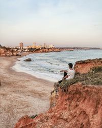 Rear view of man sitting on rock by sea against sky